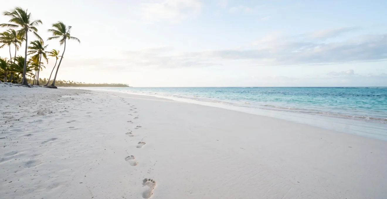 Wide editorial view of a pristine white-sand stretch of Bavaro Beach with bare footprints leading toward turquoise water at sunrise